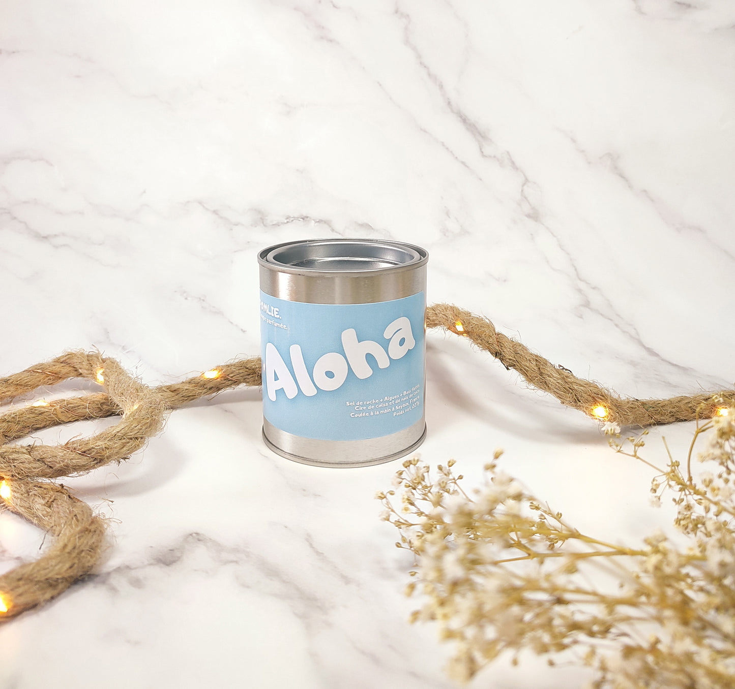 La bougie parfumée Aloha fermée qui côtoie une corde de décoration et quelques fleurs séchées.