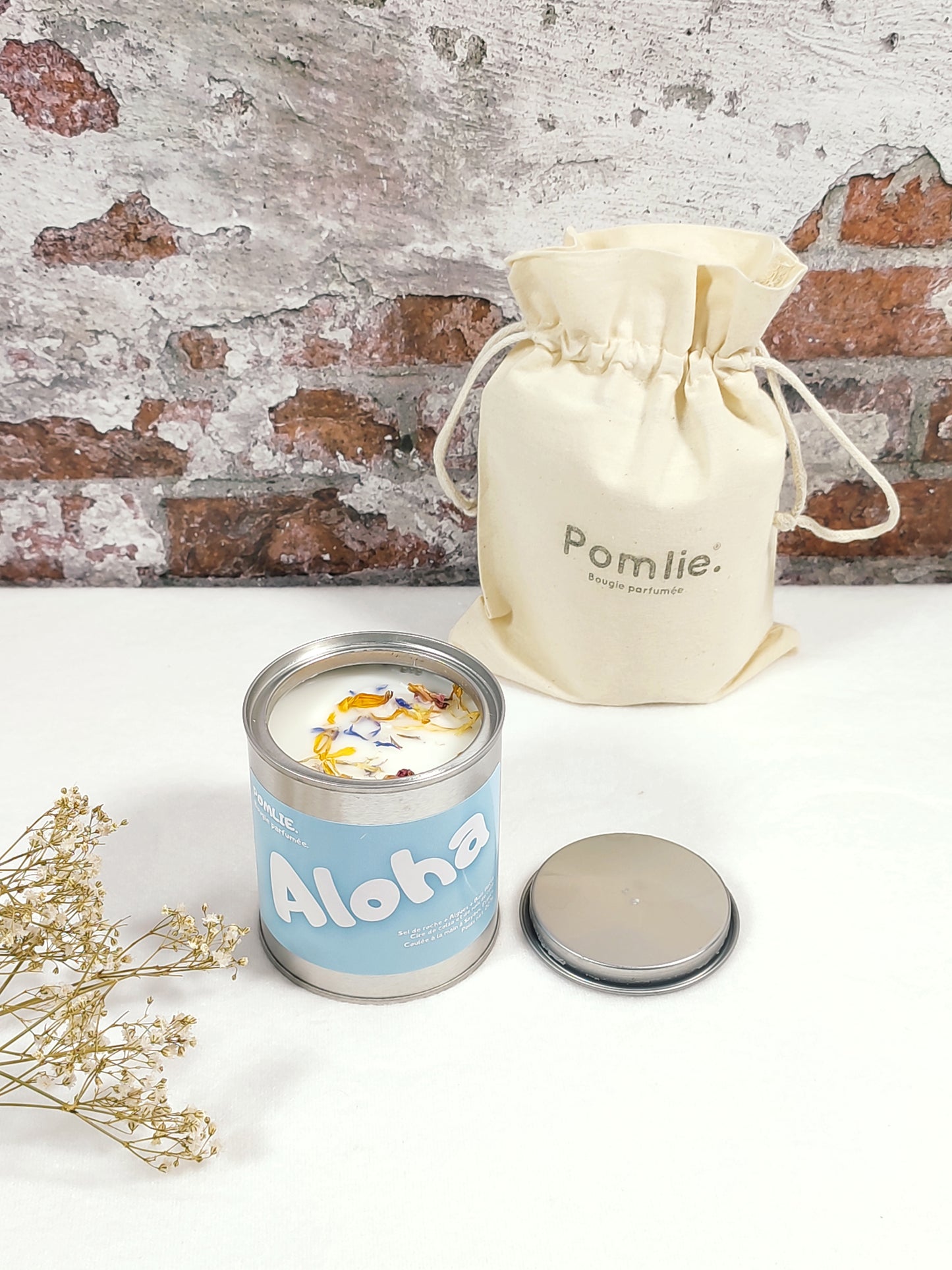 Cette bougie Pomlie est présentée dans une petite boîte en métal bleue avec l’inscription ‘Aloha’ avec à coté d'elle sont sac en coton.