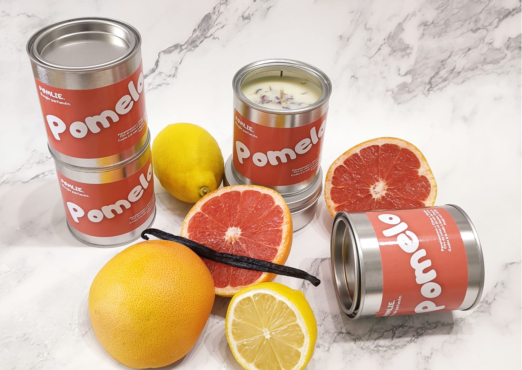 Bougie Pomlie Pomelo en métal rouge avec pamplemousse, citron et vanille à ces côtés.